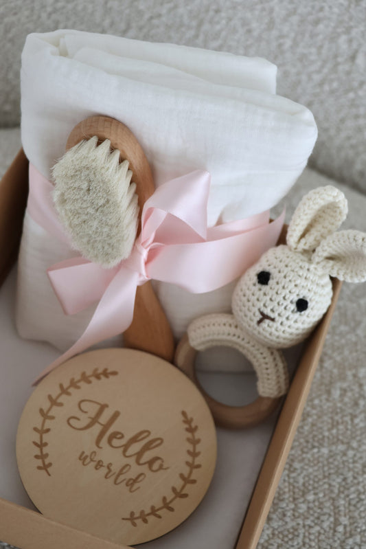 Coffret cadeau naissance pour bébé en bois et coton sur fond en bois, avec hochet lapin en crochet, lange tetra blanc et brosse à cheveux douce pour bébé.