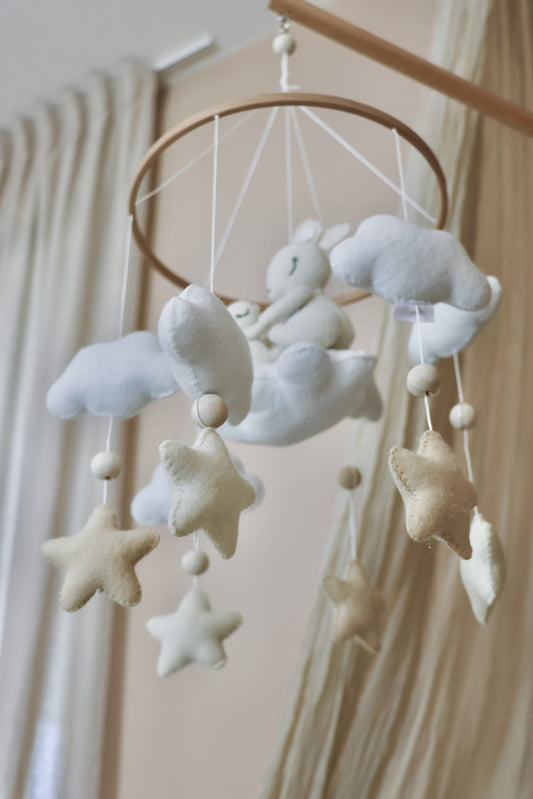 Mobile de berceau en bois pour bébé avec des peluches en feutrine blanche représentant une maman lapin et son petit assis sur un nuage, entourés d'étoiles beige et de nuages blancs.