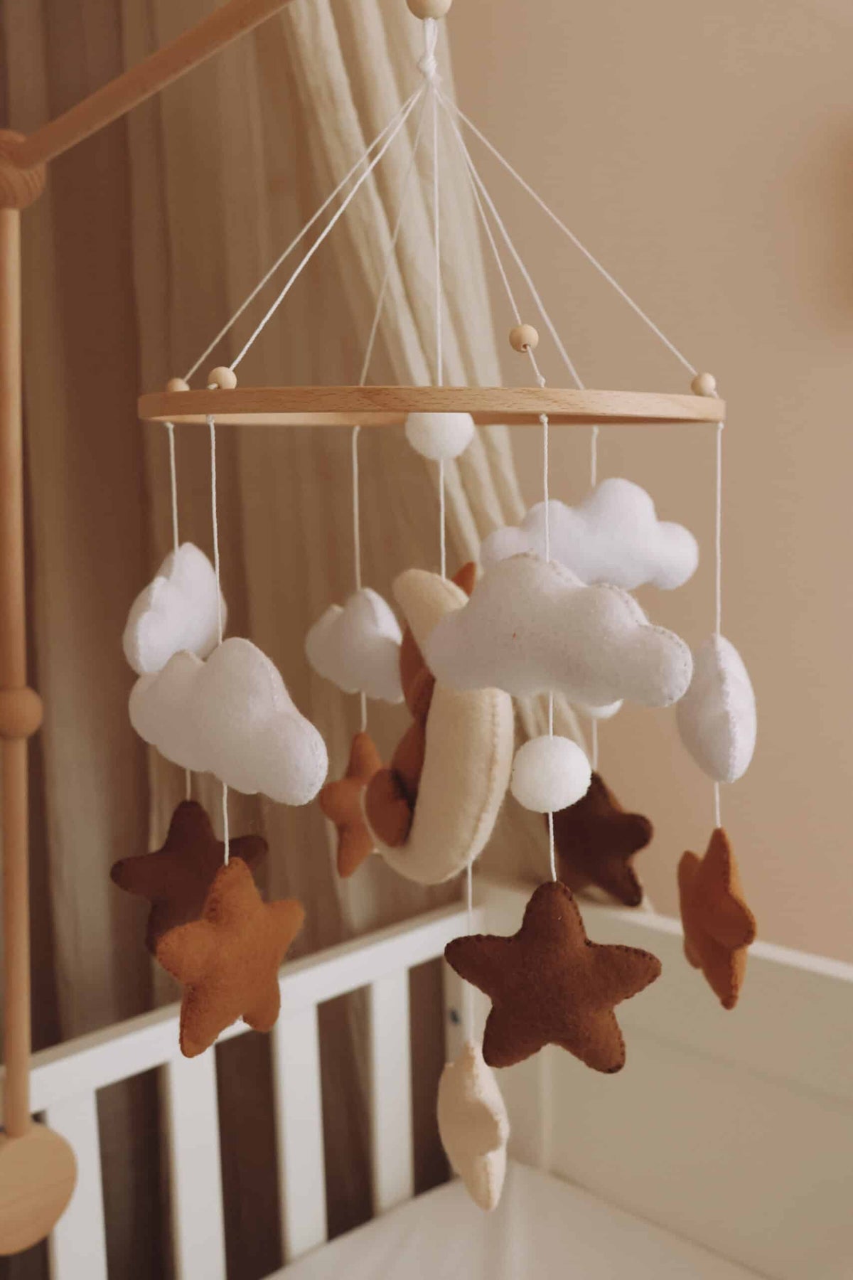 Mobile artisanal pour berceau avec un petit ours brun endormi sur une lune blanche en peluche entouré de nuages blancs, suspendu au dessus des étoiles en tissu beige, marron et blanc.