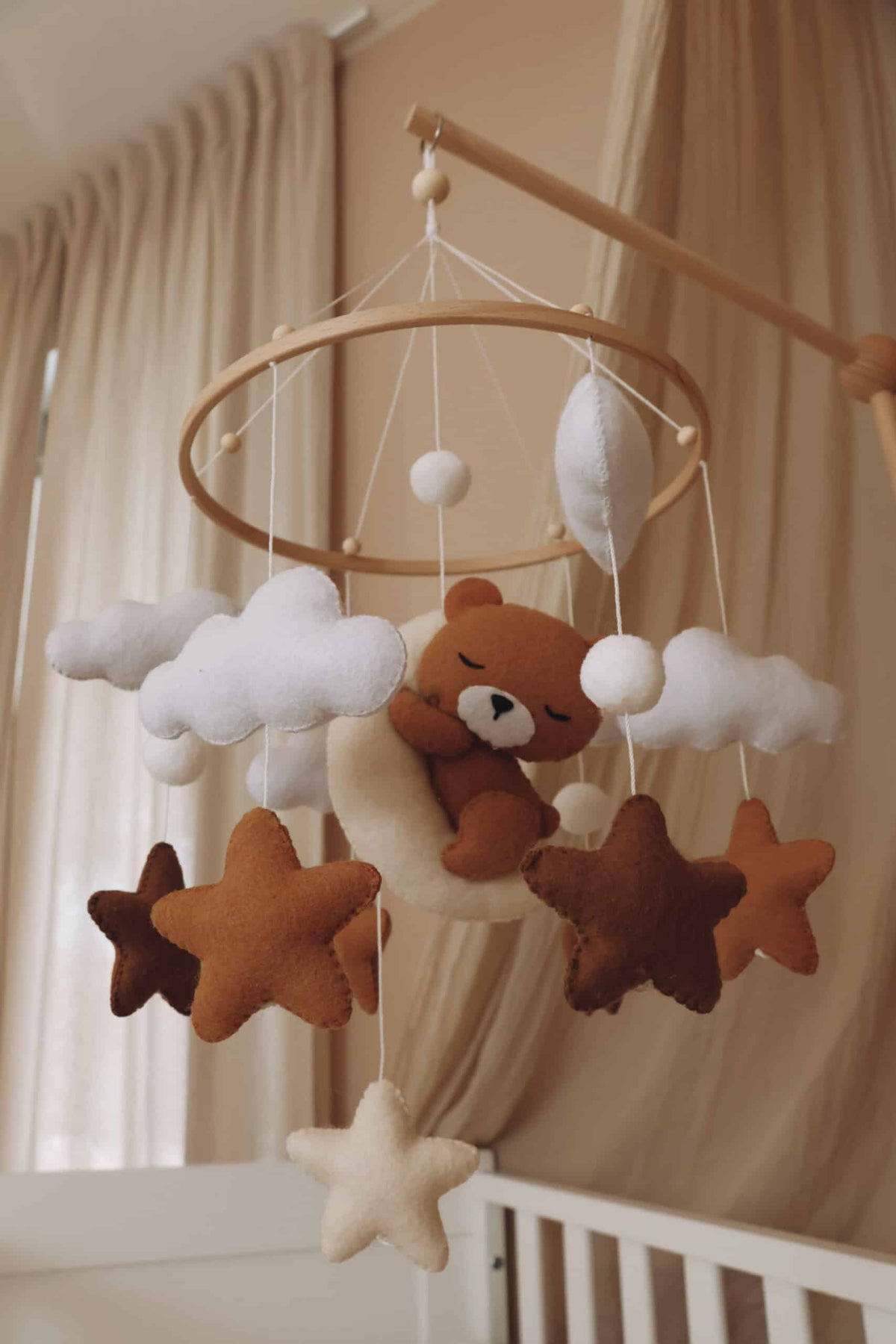 Mobile artisanal pour berceau avec un petit ours brun endormi sur une lune blanche en peluche entouré de nuages blancs, suspendu au dessus des étoiles en tissu beige, marron et blanc.