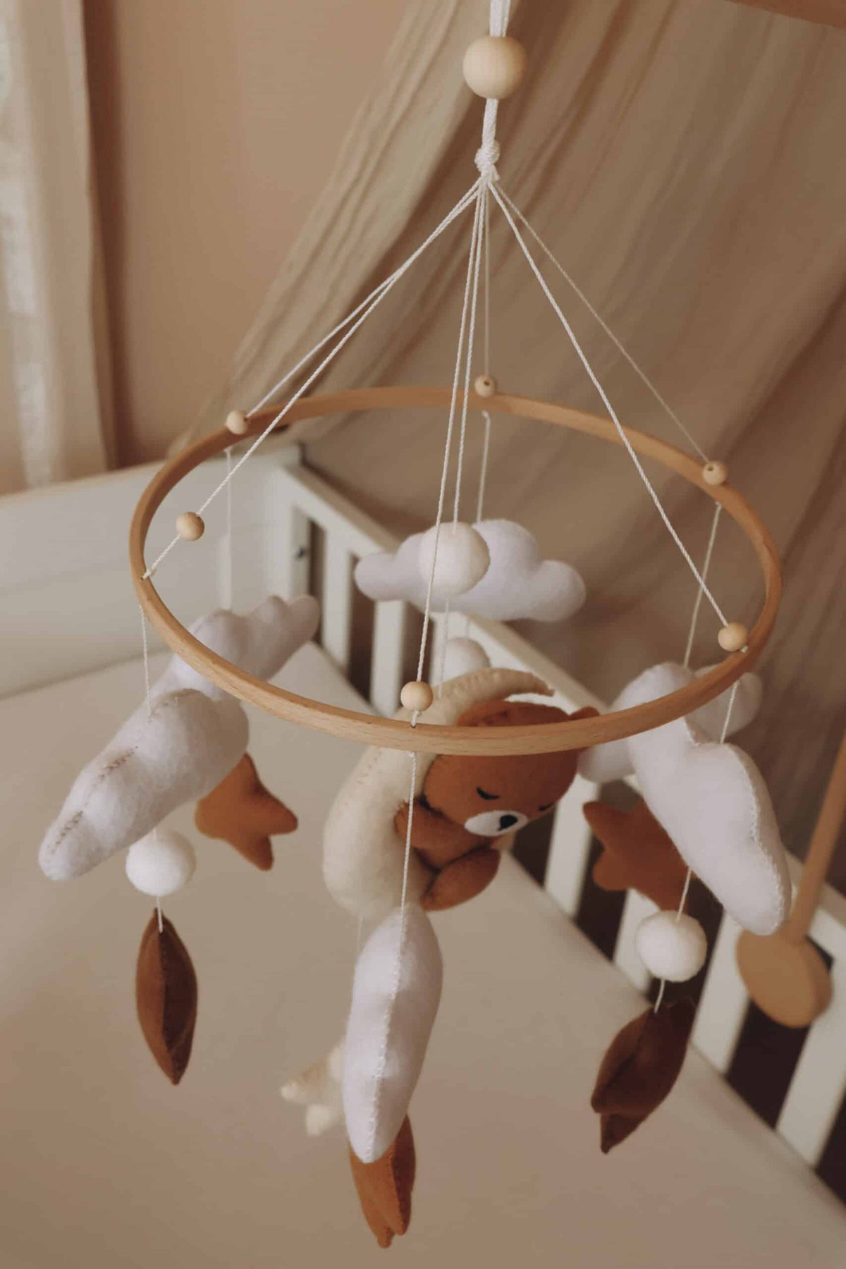 Mobile artisanal pour berceau avec un petit ours brun endormi sur une lune blanche en peluche entouré de nuages blancs, suspendu au dessus des étoiles en tissu beige, marron et blanc.