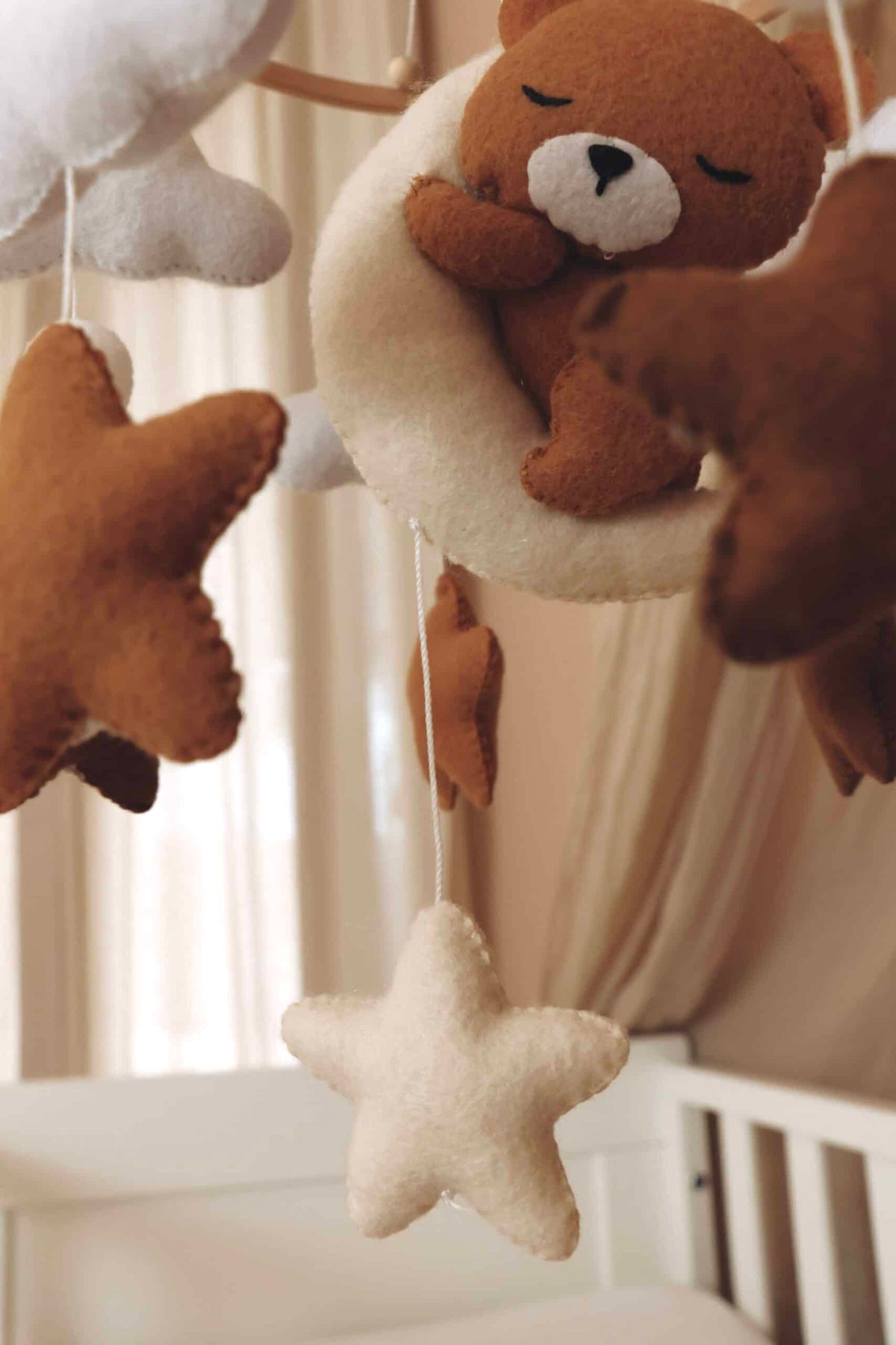 Mobile artisanal pour berceau avec un petit ours brun endormi sur une lune blanche en peluche entouré de nuages blancs, suspendu au dessus des étoiles en tissu beige, marron et blanc. détails vus de près