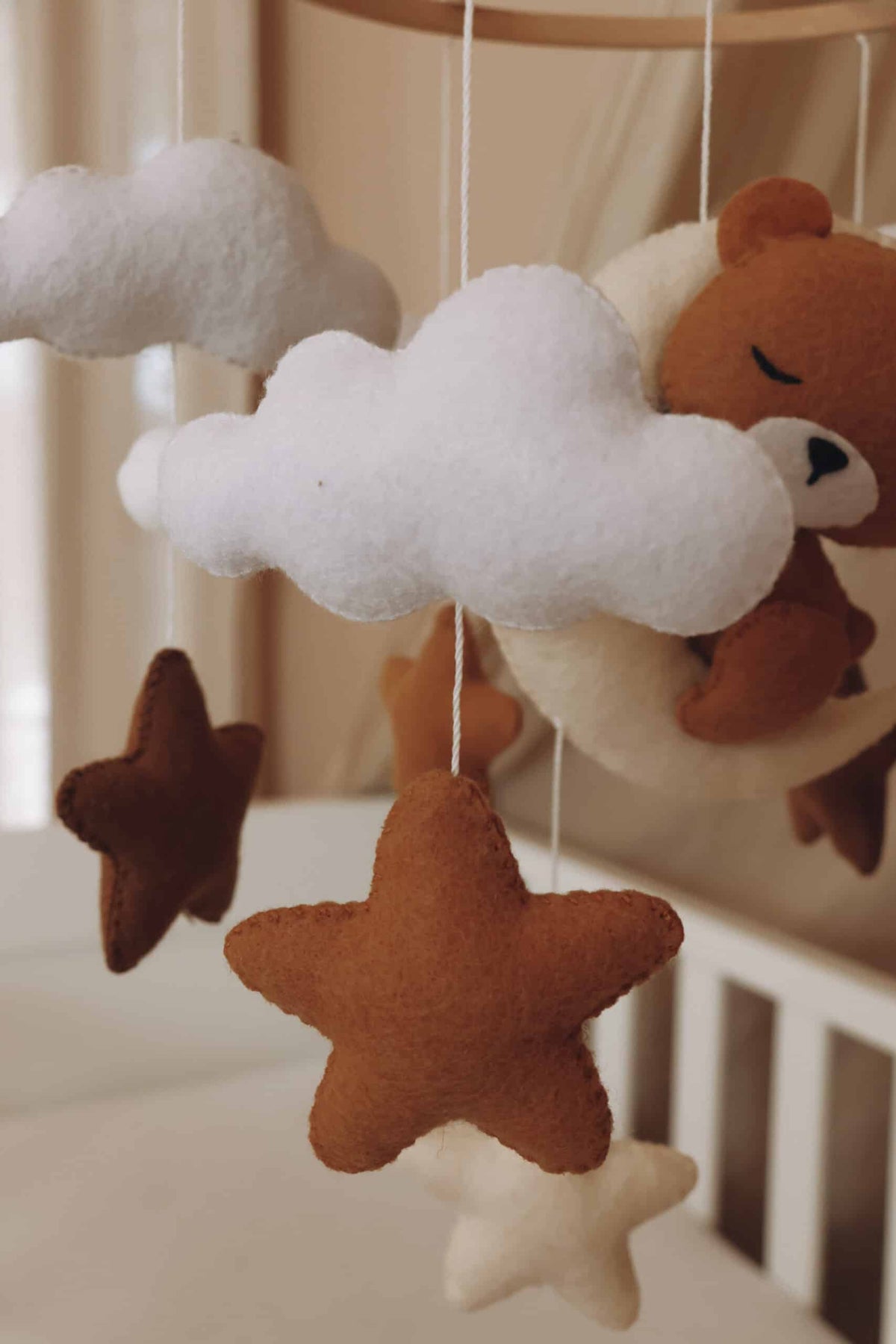 Mobile artisanal pour berceau avec un petit ours brun endormi sur une lune blanche en peluche entouré de nuages blancs, suspendu au dessus des étoiles en tissu beige, marron et blanc. détails vus de près