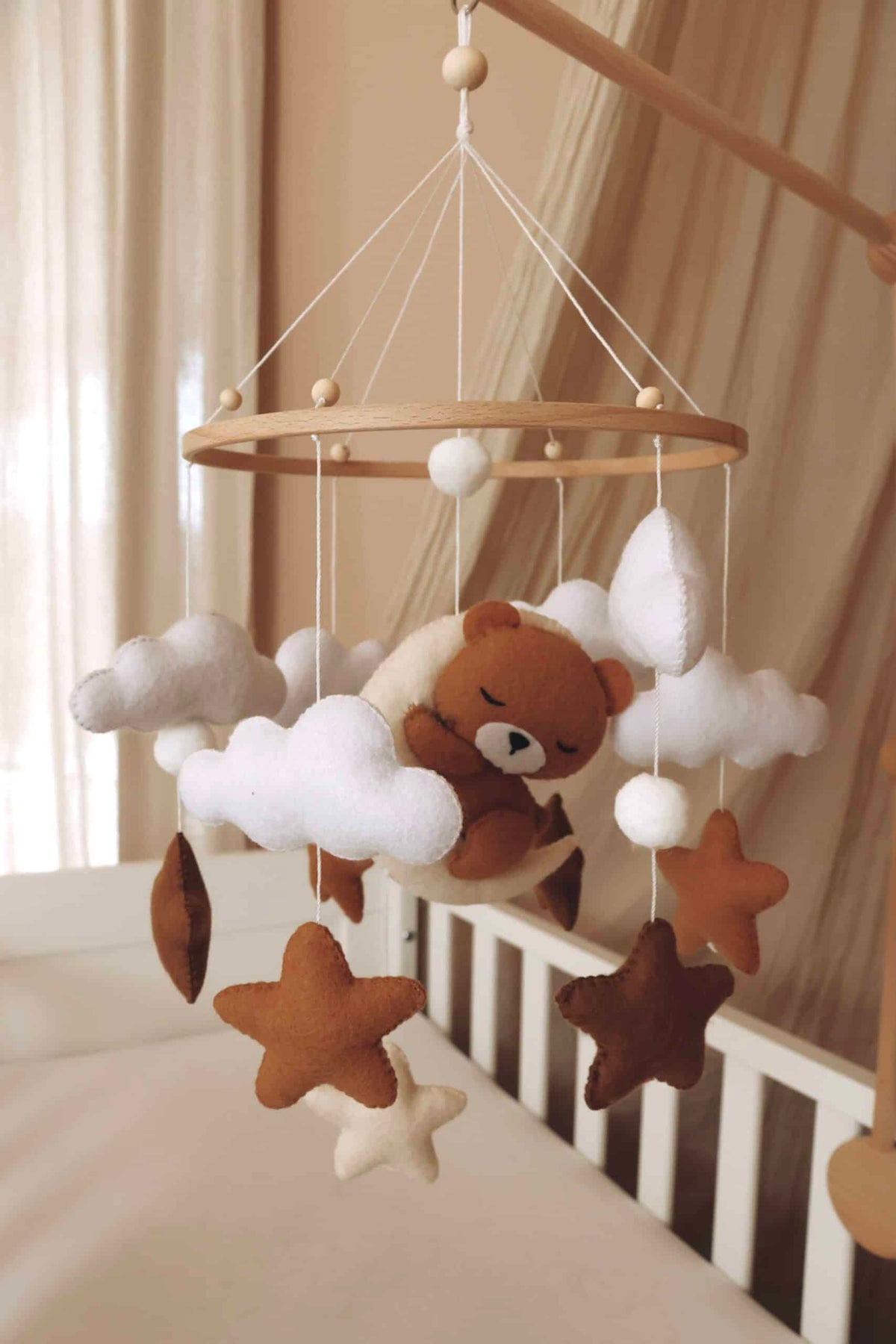 Mobile artisanal pour berceau avec un petit ours brun endormi sur une lune blanche en peluche entouré de nuages blancs, suspendu au dessus des étoiles en tissu beige, marron et blanc.
