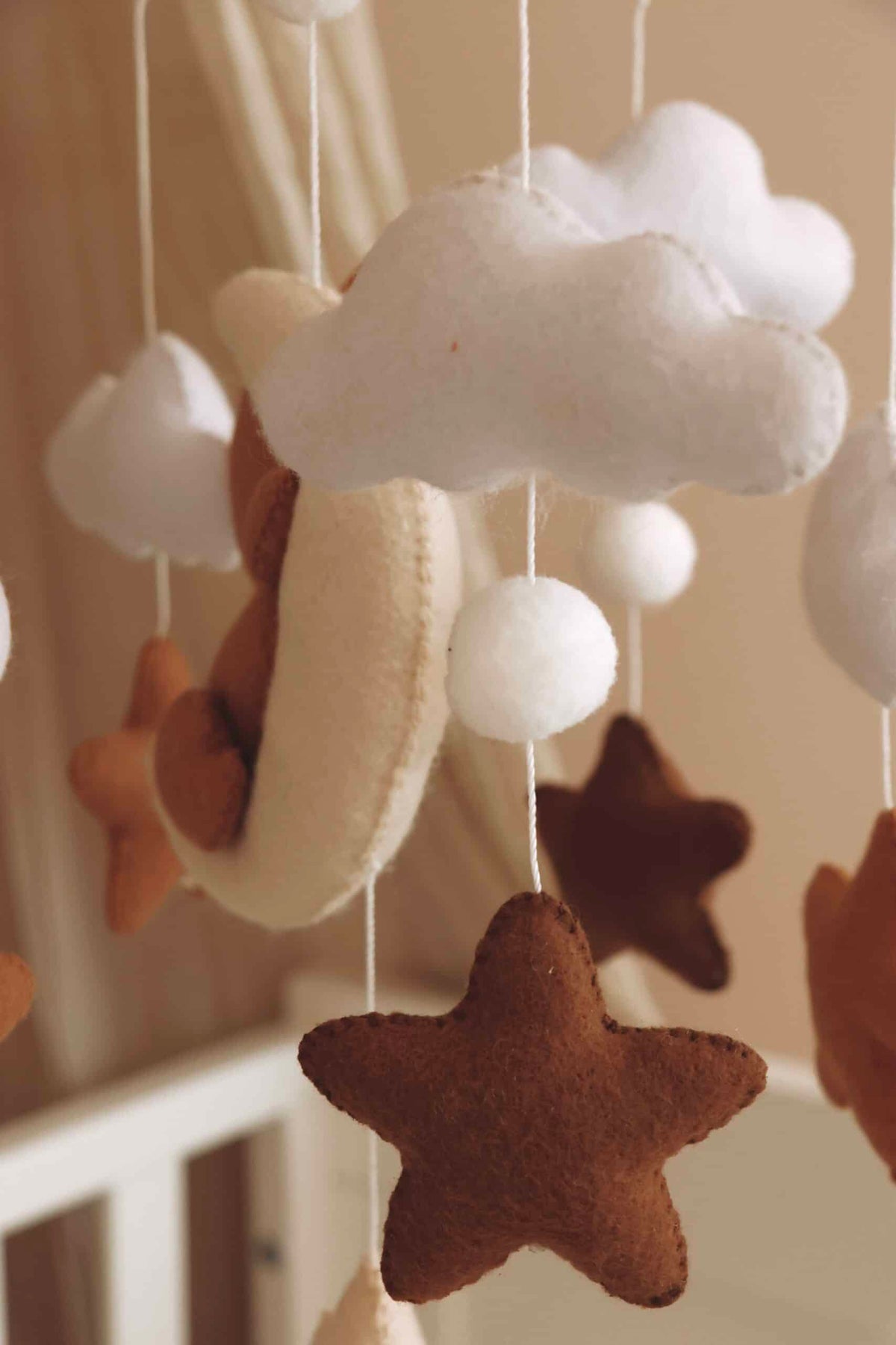 Vue de près. Mobile artisanal pour berceau avec un petit ours brun endormi sur une lune blanche en peluche entouré de nuages blancs, suspendu au dessus des étoiles en tissu beige, marron et blanc.