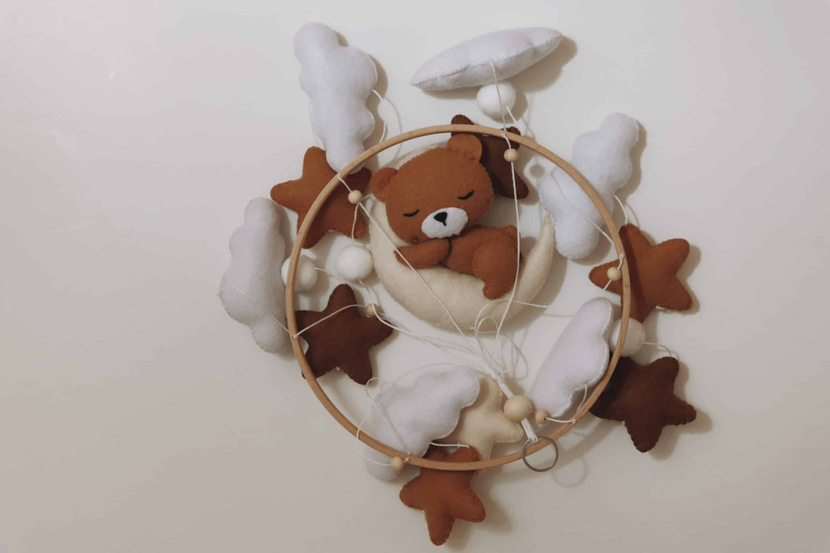 Mobile artisanal pour berceau avec un petit ours brun endormi sur une lune blanche en peluche entouré de nuages blancs, suspendu au dessus des étoiles en tissu beige, marron et blanc. non suspendu