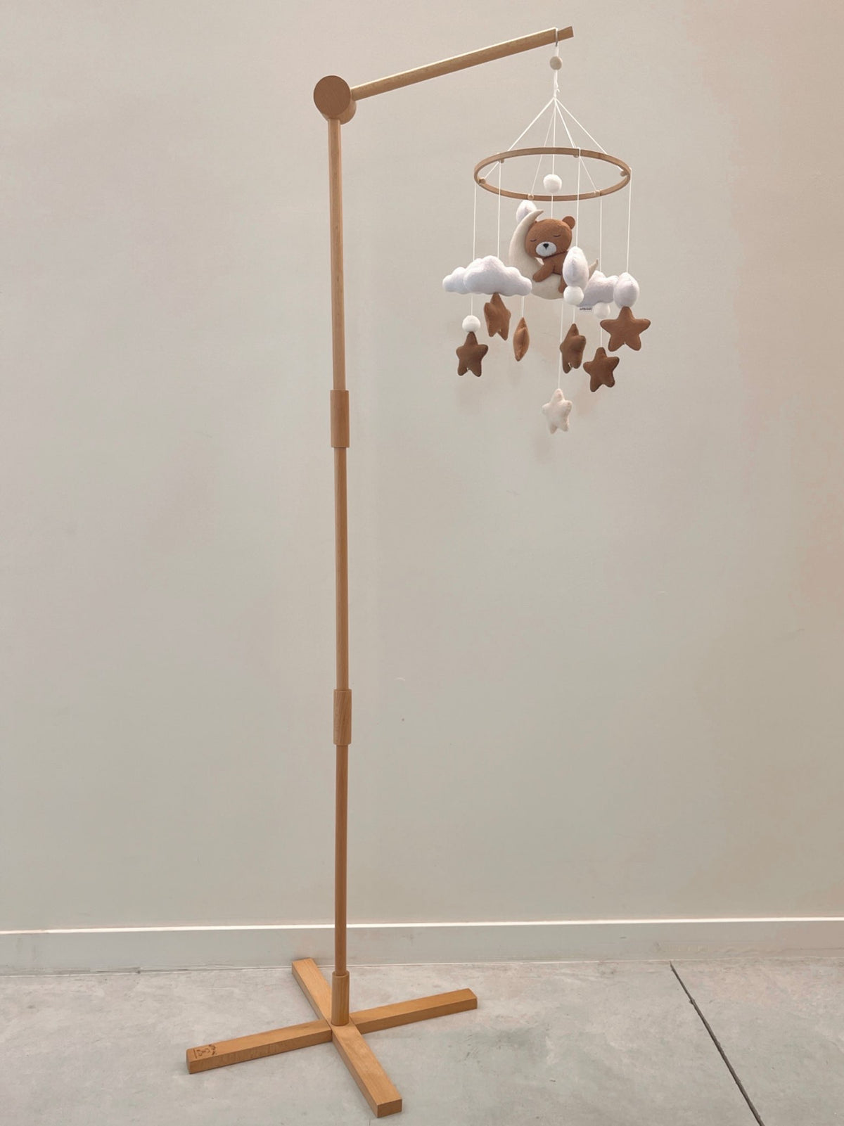 Présentation du mobile et de sa potence en bois. Mobile artisanal pour berceau avec un petit ours brun endormi sur une lune blanche en peluche entouré de nuages blancs, suspendu au dessus des étoiles en tissu beige, marron et blanc. 