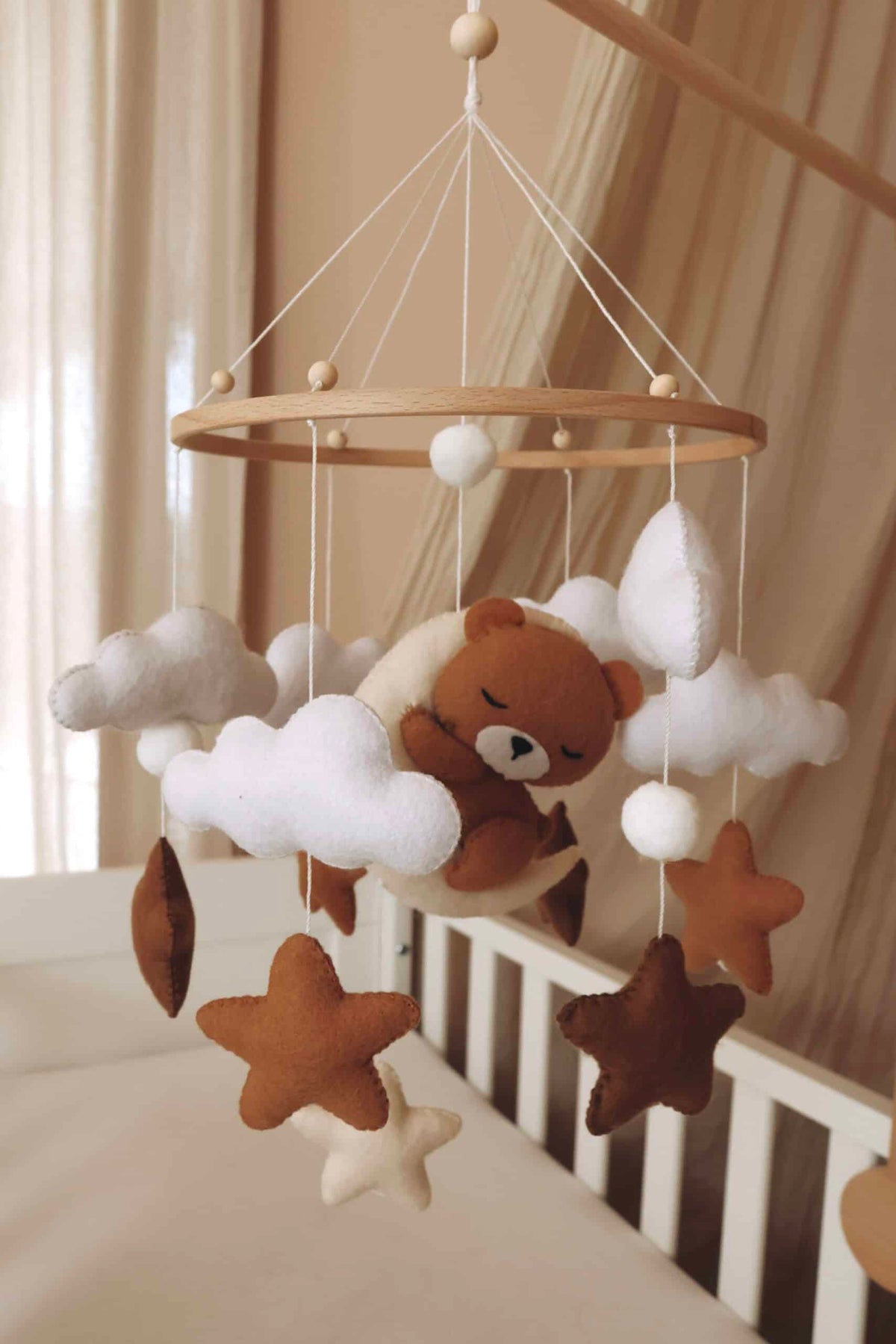 Mobile artisanal pour berceau avec un petit ours brun endormi sur une lune blanche en peluche entouré de nuages blancs, suspendu au dessus des étoiles en tissu beige, marron et blanc.