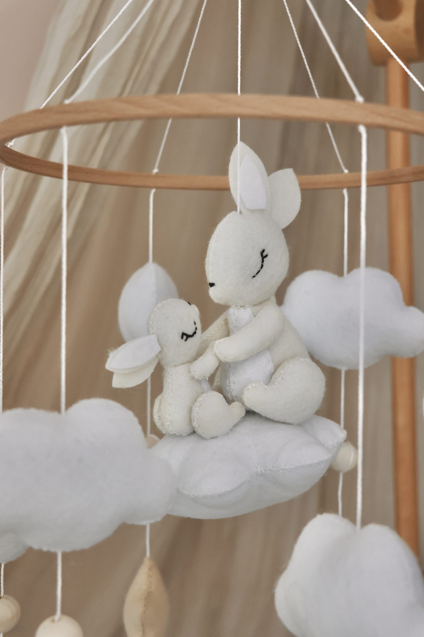 Mobile de berceau en bois pour bébé avec des peluches en feutrine blanche représentant une maman lapin et son petit assis sur un nuage, entourés d'étoiles beige et de nuages blancs.