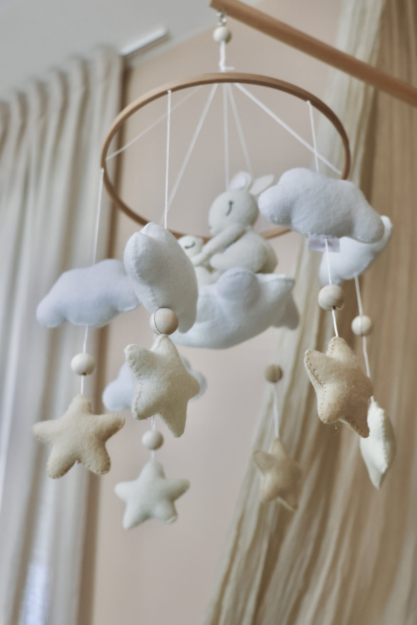 Mobile de berceau en bois pour bébé avec des peluches en feutrine blanche représentant une maman lapin et son petit assis sur un nuage, entourés d'étoiles beige et de nuages blancs.