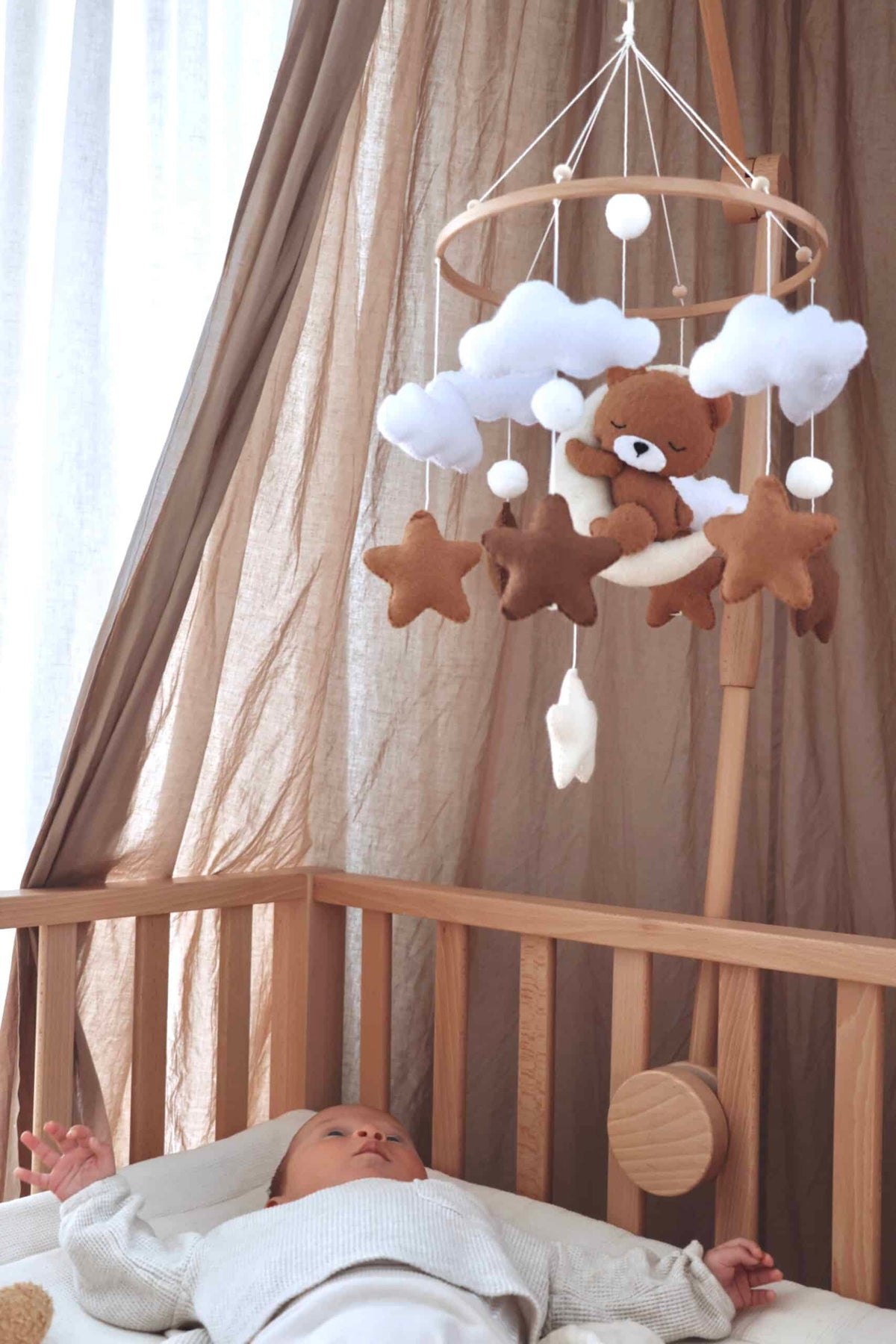Mobile artisanal pour berceau avec un petit ours brun endormi sur une lune blanche en peluche entouré de nuages blancs, suspendu au dessus des étoiles en tissu beige, marron et blanc.