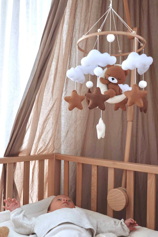 Mobile artisanal pour berceau avec un petit ours brun endormi sur une lune blanche en peluche entouré de nuages blancs, suspendu au dessus des étoiles en tissu beige, marron et blanc.